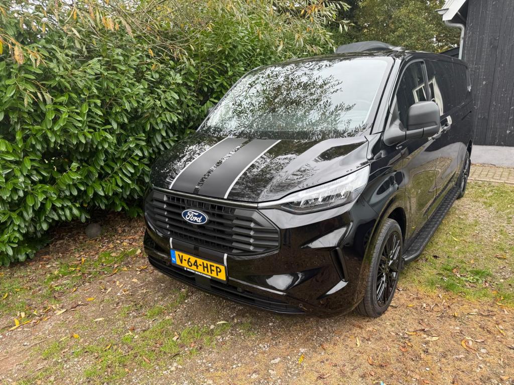 Een zwarte Ford Transit Custom geparkeerd op een grasachtige ondergrond, met een bushouding en groene struiken op de achtergrond.