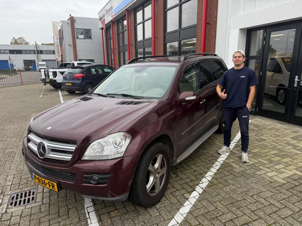 Een man staat naast een maroon Mercedes SUV, met een 'thumbs up'-gebaar, voor een showroom met andere auto's op de achtergrond.